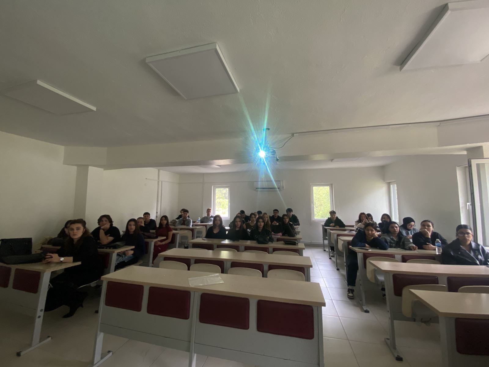 In a bright classroom, approximately 20–25 university students are seated at desks, looking toward the camera. The classroom is simple and orderly, with white walls, light-colored desks, and rectangular LED lights on the ceiling. A projector light hanging from the ceiling is visible in the center of the room. The students appear to be young adults, mostly seated and attentive. There are windows at the back of the classroom, allowing natural light to enter.