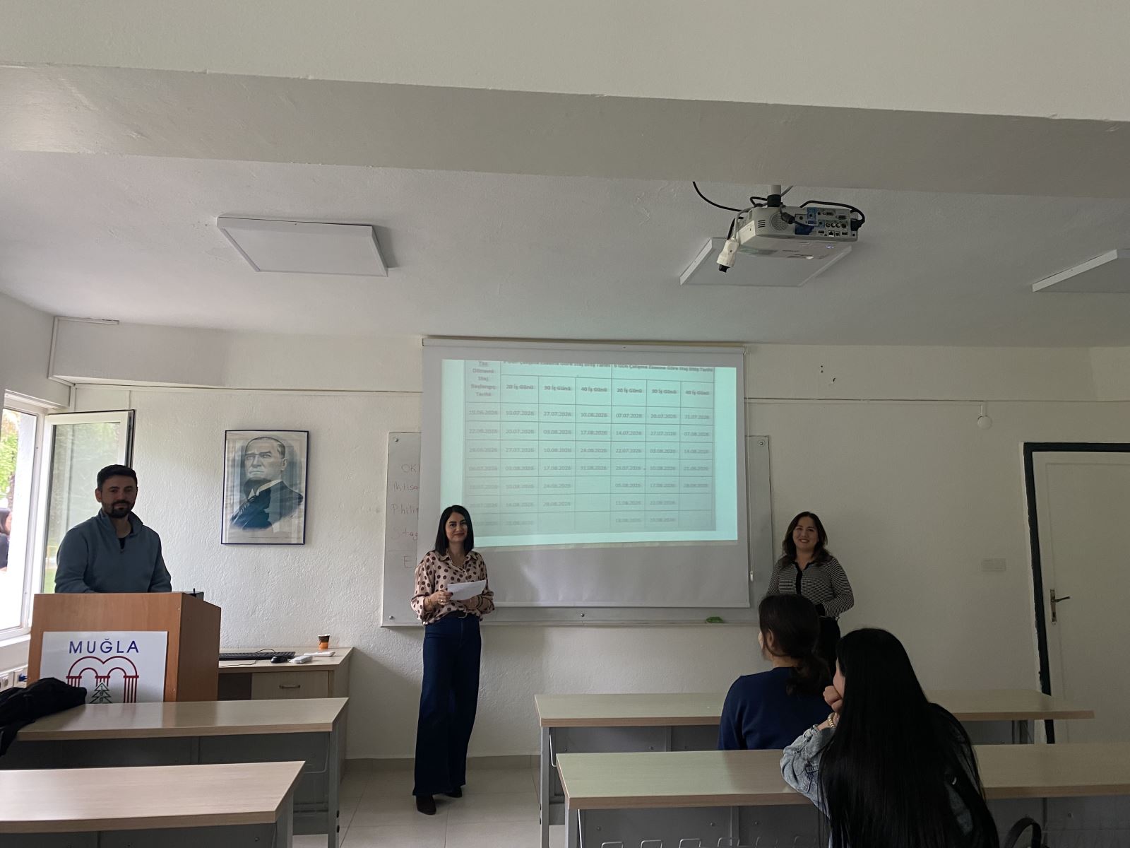 At the front of a classroom, three academic staff members are standing. On the left, a male lecturer stands behind a podium. In the center, a female lecturer holds a piece of paper, and on the right, another female lecturer stands beside her. Behind them, a projected presentation screen displays tabular information. A portrait of Mustafa Kemal Atatürk is visible on the wall. A few students are seated in the front rows, listening to the lecturers. The classroom appears bright and well-organized.